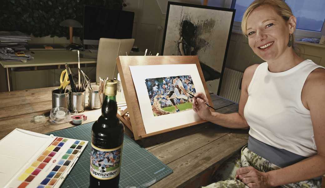 Eva Wrann bei der Arbeit zu Hause in einem Zimmer, das sie als Atelier eingerichtet hat. Nebst dem Originalbild auf kartoniertem A3-Blatt ist auch eine «Appenzeller Alpenbitter»-Flasche mit dem Werk auf der Etikette zu sehen.  
