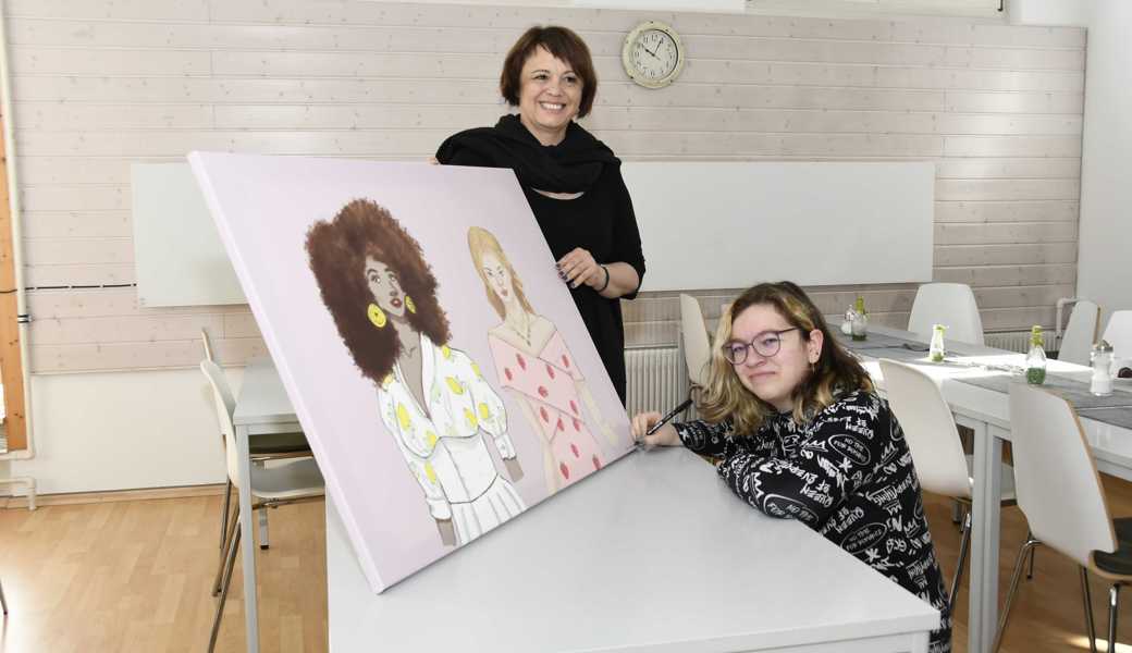 Laura Bauer (Teilnehmerin im Motivationssemester, rechts) signiert ihr Bild und übergibt es Maria Garcia (Leiterin des A-Treffs). Die dargestellten Frauen versinnbildlichen die Vielfalt der Gäste, die farbigen Kleider Freundlichkeit.